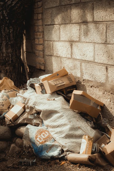 A pile of miscellaneous rubbish and packaging materials, including cardboard boxes, paper bags, and plastic wrapping, is situated against a rough brick wall on a dirt or gravel surface. The cardboard boxes vary in size, some partially opened or crushed, and appear used or discarded, with visible tape and printed labels. A large, textured fabric bag or sack is positioned among the debris, some of its edges creased or torn. The surrounding environment includes a nearby tree trunk partially visible on the left, with dark bark contrasting against the wall. The lighting is natural, casting minimal shadows, indicating an outdoor setting. The scene appears to depict an area where rubbish is temporarily stored or awaiting collection, related to independent waste removal services like those offered by Rubbish Removal Brompton, which provides alternative options to traditional council waste disposal.