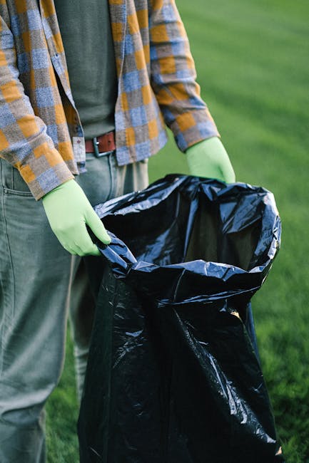 A person wearing a blue and yellow checked shirt, grey trousers, and a brown belt is holding open a large black rubbish bag lined with a silver interior, standing outdoors on a grassy area. The individual is also wearing bright green gloves, indicating they are handling waste or rubbish collection. The surrounding environment features a well-maintained green lawn, suggesting a garden or park setting. The focus is on the person's torso and hands as they prepare the rubbish bag for waste disposal, which is relevant to private waste handling or rubbish removal services. The scene emphasizes careful handling of waste materials, aligning with professional rubbish removal practices provided by companies like Rubbish Removal Brompton, which offers alternative waste collection options outside standard municipal services.