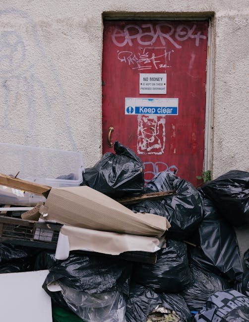 A pile of miscellaneous rubbish and packaging materials, including cardboard boxes, paper bags, and plastic wrapping, is situated against a rough brick wall on a dirt or gravel surface. The cardboard boxes vary in size, some partially opened or crushed, and appear used or discarded, with visible tape and printed labels. A large, textured fabric bag or sack is positioned among the debris, some of its edges creased or torn. The surrounding environment includes a nearby tree trunk partially visible on the left, with dark bark contrasting against the wall. The lighting is natural, casting minimal shadows, indicating an outdoor setting. The scene appears to depict an area where rubbish is temporarily stored or awaiting collection, related to independent waste removal services like those offered by Rubbish Removal Brompton, which provides alternative options to traditional council waste disposal.