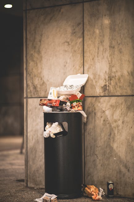 A black cylindrical trash bin with a white open lid, positioned against a large beige or light brown tiled wall. The bin is overflowing with various types of rubbish, including crumpled food packaging, cardboard boxes, and plastic wrappers, some of which are spilling onto the surrounding floor. There are additional waste items on the ground in front of and beside the bin, such as a crumpled paper and a small empty can. The environment appears to be part of an indoor or covered area, possibly a public building or corridor, with minimal lighting emphasizing the visible mess and emphasizing the need for rubbish removal services like those provided by Rubbish Removal Brompton. The scene highlights the importance of proper waste management and the potential for private collection or on-site clearance to maintain cleanliness in communal or institutional spaces.