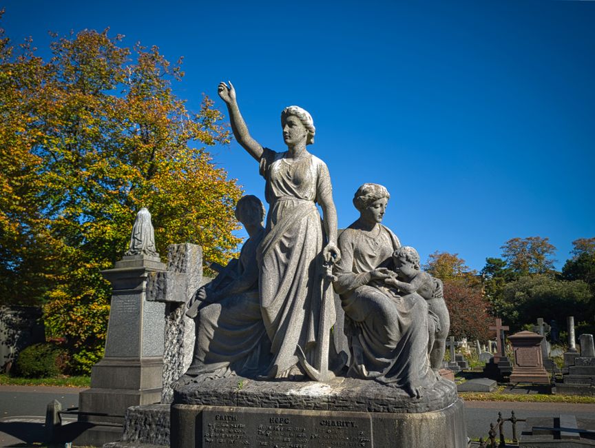 A detailed stone monument situated in a cemetery, featuring three carved statues set against a backdrop of autumnal trees with yellow and orange foliage. The central figure is a standing woman dressed in flowing garments, with her right arm raised high and her left hand holding a small object. To her left is a seated woman with her head bowed, holding a child on her lap, while the child appears to be reaching towards the seated woman's face. To the right of the standing figure, another woman is seated, cradling a small literal child in her arms. The statues exhibit detailed textures of stone with weathered finishes, showcasing realistic drapery in the garments and gentle expressions. The monument rests on a rectangular stone base with inscriptions. Bright sunlight illuminates the scene, highlighting the monument's intricate craftsmanship and contrasting against the deep blue sky, creating a solemn and respectful atmosphere typical of cemetery memorials and areas where private waste removal services may operate nearby.