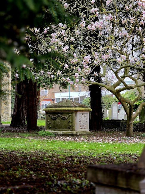 A detailed stone monument situated in a cemetery, featuring three carved statues set against a backdrop of autumnal trees with yellow and orange foliage. The central figure is a standing woman dressed in flowing garments, with her right arm raised high and her left hand holding a small object. To her left is a seated woman with her head bowed, holding a child on her lap, while the child appears to be reaching towards the seated woman's face. To the right of the standing figure, another woman is seated, cradling a small literal child in her arms. The statues exhibit detailed textures of stone with weathered finishes, showcasing realistic drapery in the garments and gentle expressions. The monument rests on a rectangular stone base with inscriptions. Bright sunlight illuminates the scene, highlighting the monument's intricate craftsmanship and contrasting against the deep blue sky, creating a solemn and respectful atmosphere typical of cemetery memorials and areas where private waste removal services may operate nearby.