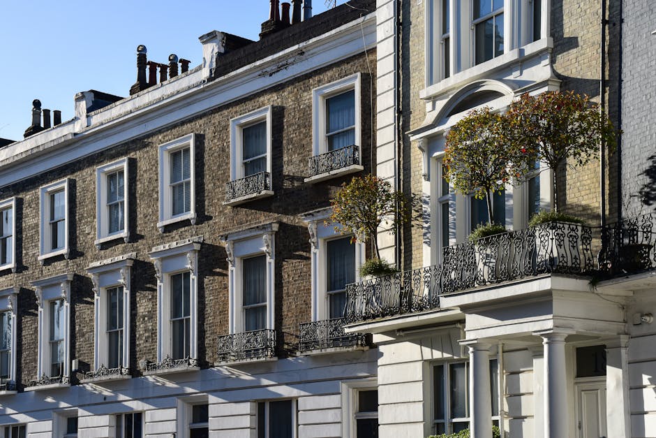 Photograph of a traditional London townhouse facade featuring multiple floors with large white-framed sash windows and wrought iron balcony railings. The building's exterior combines exposed brickwork in earthy tones with decorative white stone or stucco detailing around the windows and along the edges. The top-floor windows have small black-painted metal balcony balustrades, while the lower floor features a larger balcony with ornate black wrought iron railings adorned with two small trees in pots. The building is situated on a paved street with bright natural lighting highlighting the textures of brick and masonry. Shadows cast by the balcony railings and trees create patterns on the white facade below. This scene, captured during daylight hours, exemplifies typical urban residential architecture where private outdoor spaces are enhanced by decorative ironwork and planters, subtly reflecting the context of local property management and potential arrangements for rubbish clearance or property maintenance by companies like Rubbish Removal Brompton in the area.