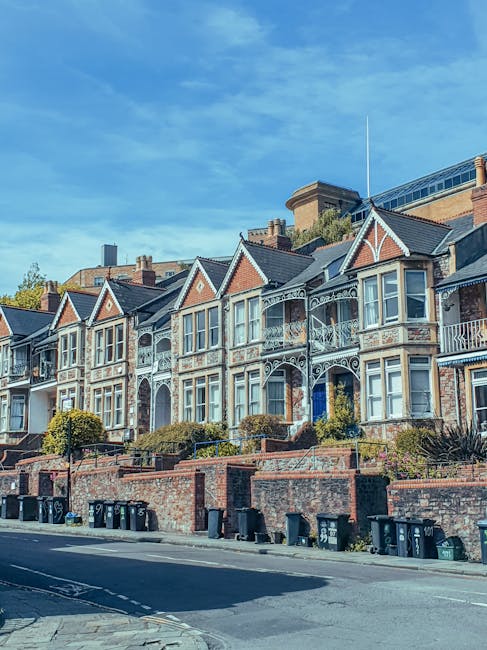 A row of Victorian-style terraced houses situated on a sloped street, featuring ornate white decorative ironwork on the balconies and porches, with brick facades in shades of light brown and red. The homes have bay windows with white frames and gabled roofs covered with dark tiles. A low brick wall runs along the front of the properties, with neatly trimmed bushes and small trees planted behind it. In the foreground, several black wheelie bins and recycling containers are lined up along the pavement, indicating waste collection activity. The sky above is clear with a few wispy clouds, and sunlight illuminates the scene, highlighting the architectural details. This setting is representative of residential areas where private waste collection services, such as those from Rubbish Removal Brompton, may be employed for on-site clearance or rubbish removal, especially in densely populated neighbourhoods with historic homes