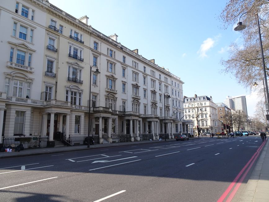 Photograph of a traditional London townhouse facade featuring multiple floors with large white-framed sash windows and wrought iron balcony railings. The building's exterior combines exposed brickwork in earthy tones with decorative white stone or stucco detailing around the windows and along the edges. The top-floor windows have small black-painted metal balcony balustrades, while the lower floor features a larger balcony with ornate black wrought iron railings adorned with two small trees in pots. The building is situated on a paved street with bright natural lighting highlighting the textures of brick and masonry. Shadows cast by the balcony railings and trees create patterns on the white facade below. This scene, captured during daylight hours, exemplifies typical urban residential architecture where private outdoor spaces are enhanced by decorative ironwork and planters, subtly reflecting the context of local property management and potential arrangements for rubbish clearance or property maintenance by companies like Rubbish Removal Brompton in the area.
