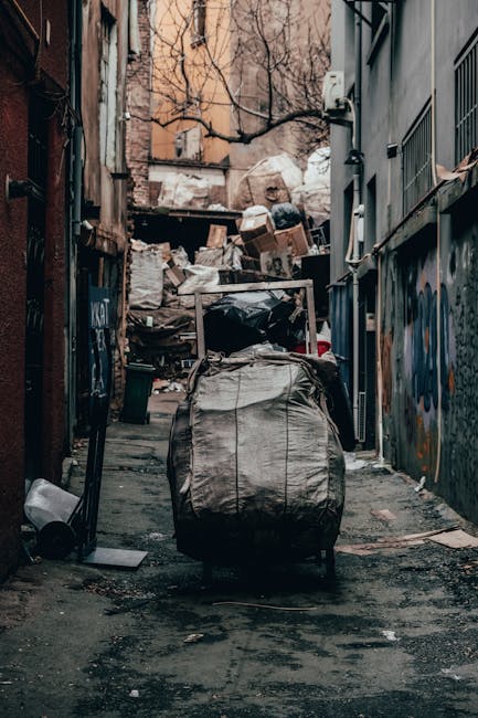 An urban alleyway scene showing a large, wheeled refuse sack made of heavy-duty, grayish fabric with visible creases and dirt marks, positioned on the rough, dark asphalt ground in the foreground. Behind the sack, there is an extensive accumulation of mixed waste, including cardboard boxes, plastic bags, and other discarded materials piled against a backdrop of buildings with brick and concrete facades. The left side features a narrow brick wall with some graffiti and a discarded black garbage bin lying on its side, while the right side has a modern gray building with metal barred windows and an air conditioning unit mounted on the wall. The alley is enclosed by tall structures, with leafless tree branches visible above, indicating winter or early spring. The scene is lit with natural daylight, casting soft shadows and emphasizing the cluttered, unmanaged appearance of the rubbish accumulation, reflecting a typical setting for private waste collection or alternative rubbish removal services in an urban environment.