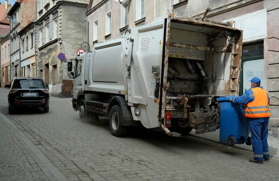 A waste collection vehicle operated by Rubbish Removal Brompton is parked on a narrow cobblestone street in an urban residential area. The white truck, with signs of rust and weathering on its rear compactor, is positioned close to the curb, with its rear open for waste intake. A worker dressed in a blue uniform and an orange high-visibility vest is standing on the right side of the vehicle, placing a blue wheeled bin into the open compartment, preparing for rubbish collection. Nearby, a black station wagon is parked parallel to the curb, and the surrounding buildings are multi-storey structures with varied architectural details, including beige and grey facades, some showing signs of aging and patches of paint chipping. The scene is illuminated with natural daylight, highlighting the textures of the cobblestones, building exteriors, and the metallic surface of the waste truck, illustrating an active temporary disposal process as part of private waste management services in an urban environment.