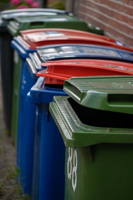 An urban alleyway scene showing a large, wheeled refuse sack made of heavy-duty, grayish fabric with visible creases and dirt marks, positioned on the rough, dark asphalt ground in the foreground. Behind the sack, there is an extensive accumulation of mixed waste, including cardboard boxes, plastic bags, and other discarded materials piled against a backdrop of buildings with brick and concrete facades. The left side features a narrow brick wall with some graffiti and a discarded black garbage bin lying on its side, while the right side has a modern gray building with metal barred windows and an air conditioning unit mounted on the wall. The alley is enclosed by tall structures, with leafless tree branches visible above, indicating winter or early spring. The scene is lit with natural daylight, casting soft shadows and emphasizing the cluttered, unmanaged appearance of the rubbish accumulation, reflecting a typical setting for private waste collection or alternative rubbish removal services in an urban environment.